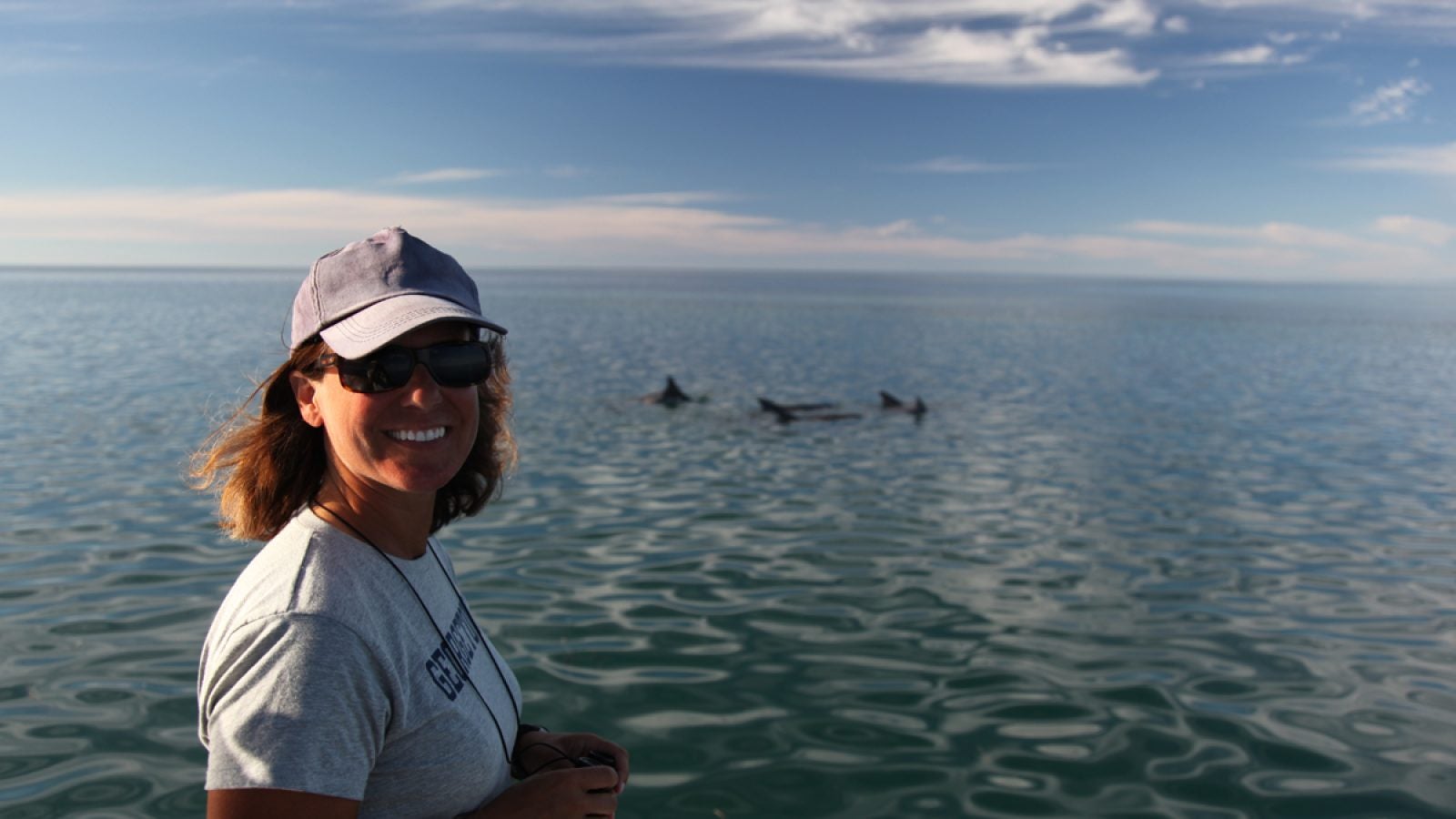 A woman in a white t shirt and hat with the ocean behind her and some dolphins in the water.