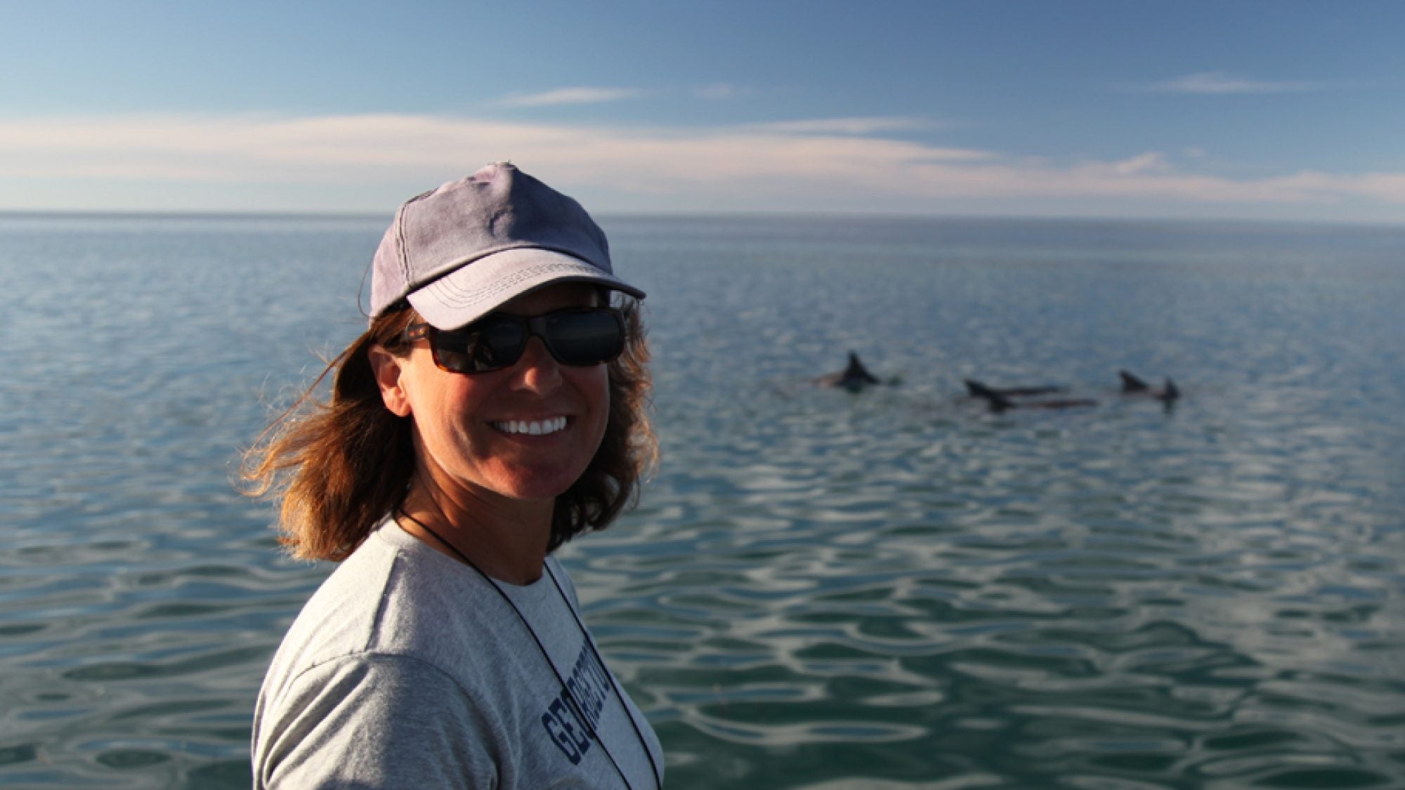 A woman in a white t shirt and hat with the ocean behind her and some dolphins in the water.