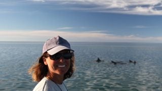 A woman in a white t shirt and hat with the ocean behind her and some dolphins in the water.