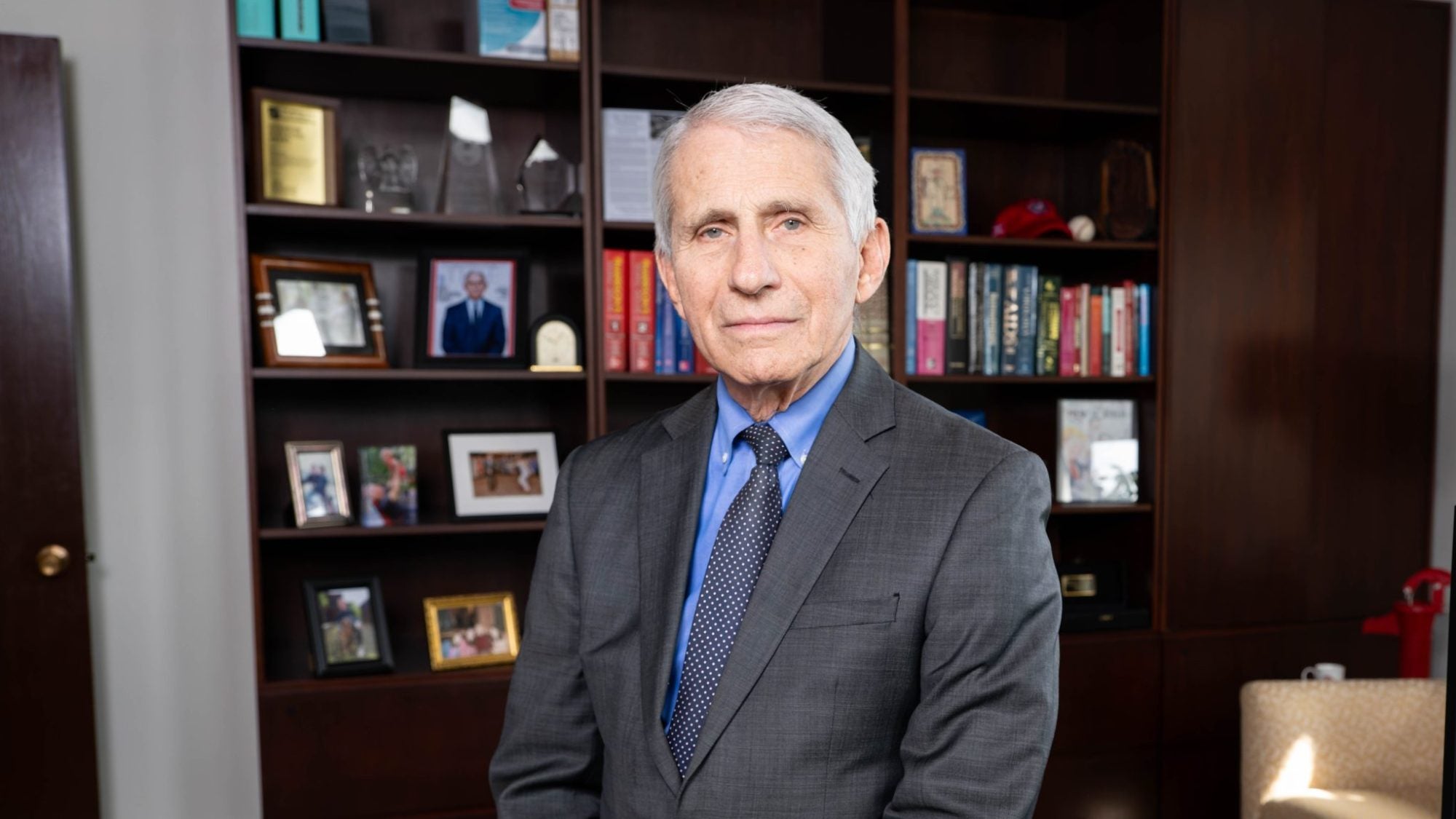Dr. Fauci leaning on a desk witth a bookcase behind him while wearing a grey suit and tie.