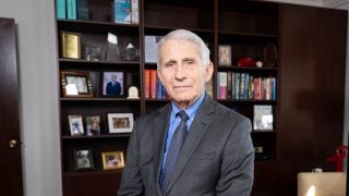 Dr. Fauci leaning on a desk witth a bookcase behind him while wearing a grey suit and tie.