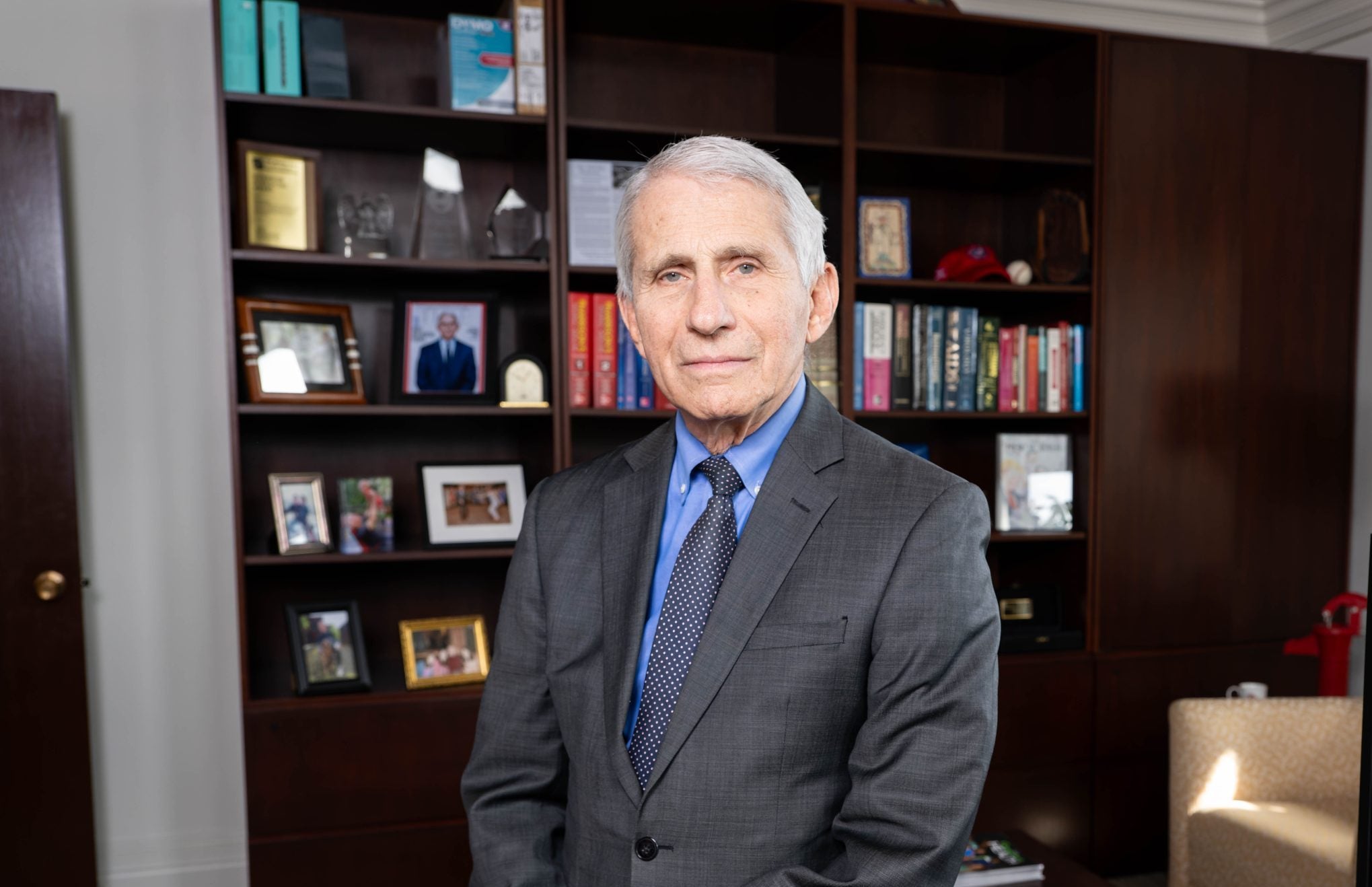 Dr. Fauci leaning on a desk witth a bookcase behind him while wearing a grey suit and tie.