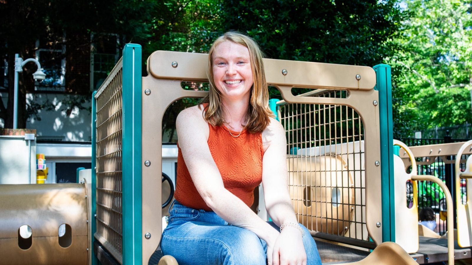 Liz Fortier sitting on a playground slide wearing jeans and an orange tank top