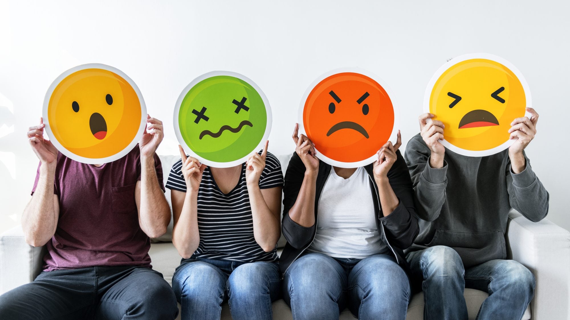 Four people sitting on a bench holding emoticon signs with different emotions