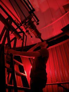 A student on a ladder peering through a telescope in the observatory in red light