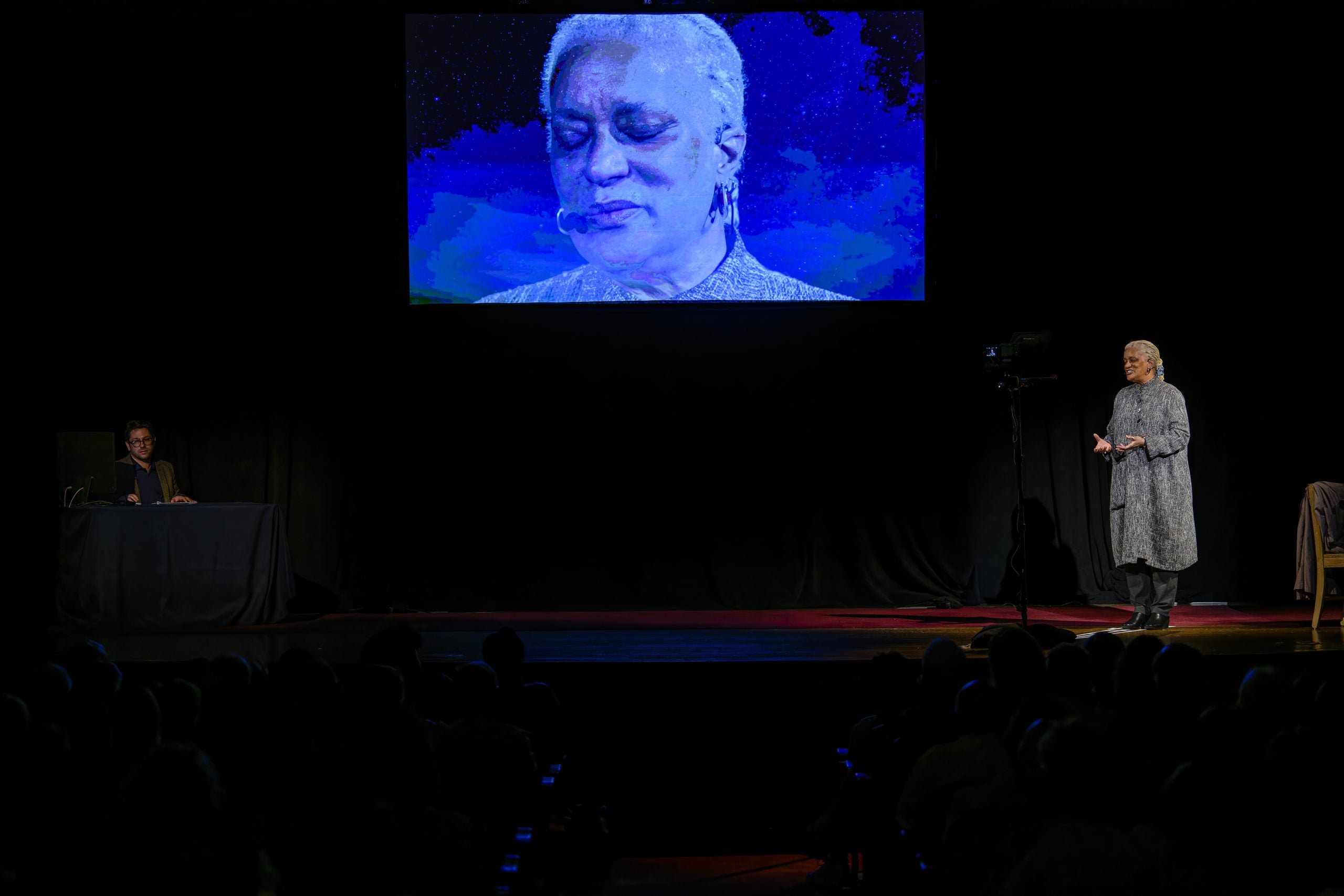 An older Black woman performing on a stage with a screen showing her face.