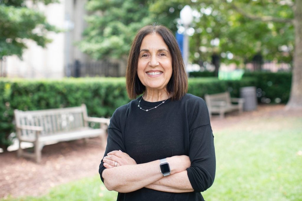 A woman with brown hair wearing a black shirt smiles with her arms folded in front of a bench.