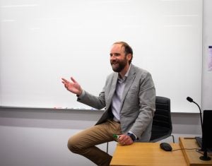 A man in a gray suit sits in a chair with one hand up smiling. A whiteboard is behind him