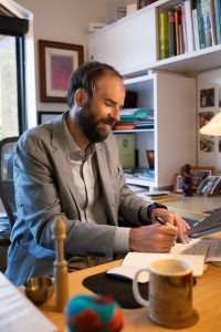 A man in a gray suit smiles as he writes in a notepad at his desk