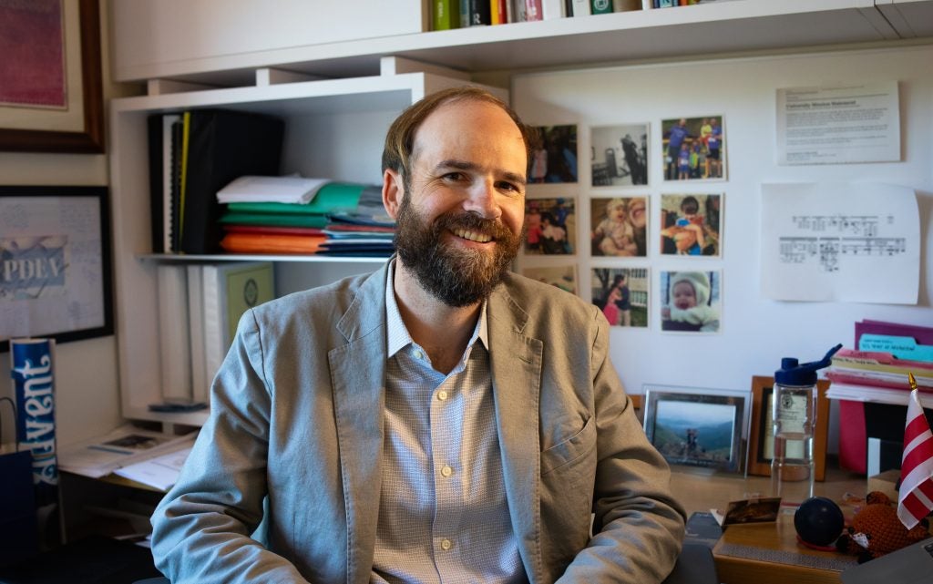 A man with brown hair and a beard smiles in a gray jacket in his office.