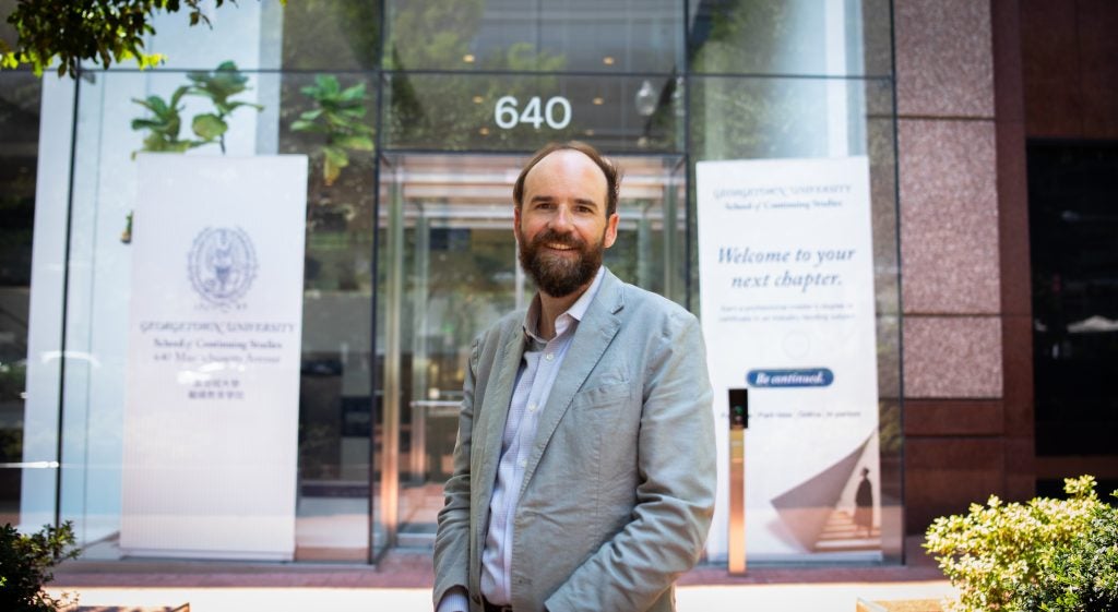 A man in a gray suit stands outside the glass entrance to Georgetown's School of Continuing Studies