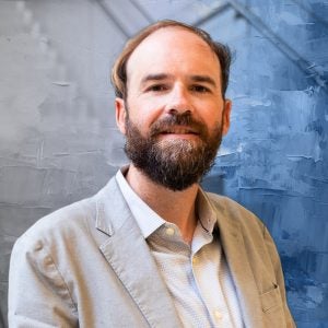 A man in a beige suit and a beard with a blue textured background behind him