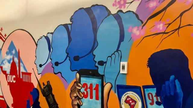 A bright orange and blue mural of silhouettes of faces with a headset on and a hand holding up a phone that says &quot;311&quot; on the screen.