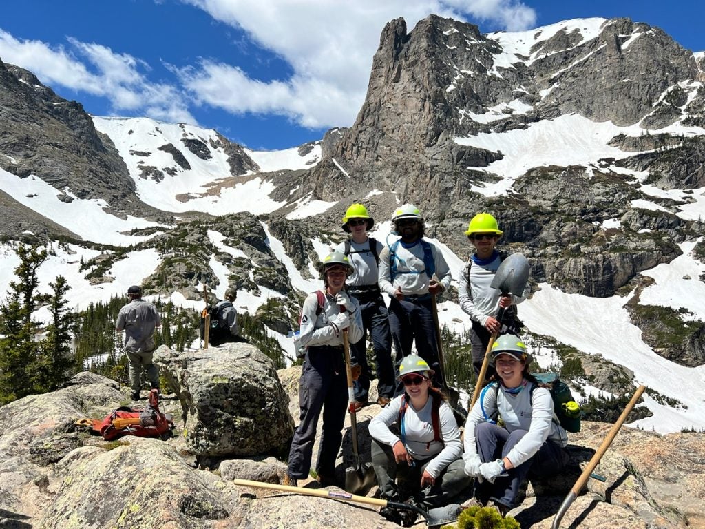 A crew of six in hard hats standing in the Rockies with some snow