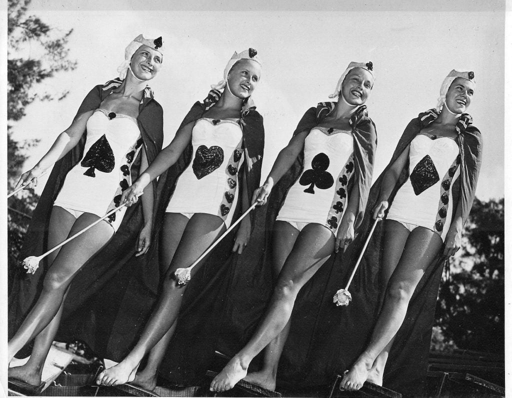 A black-and-white photo of four women in white bathing suits with an icon from a deck of cards on each of their suits. 