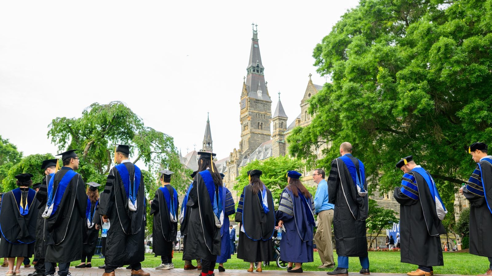 A group of people in graduation caps and gowns look up at the Georgetown clocktower.