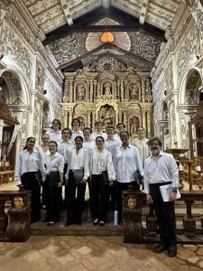 A group of Georgetown choir singers in matching white collared shirts and black pants pose together inside a church.