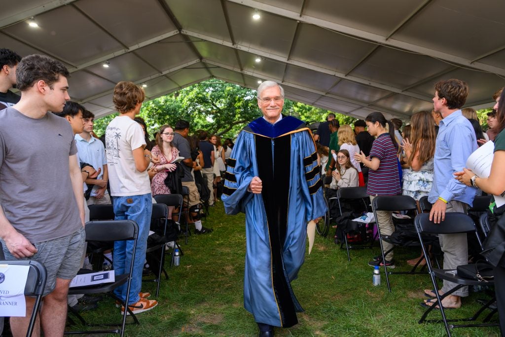 A man in a blue academic robe walks under a tent in the middle of an aisle.