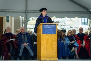 Laura DeNardis stands behind a podium in a blue and black academic robe and a black cap. 