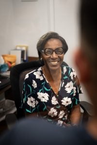 A woman with black glasses and a flowered shirt smiles at someone standing in front of the camera.