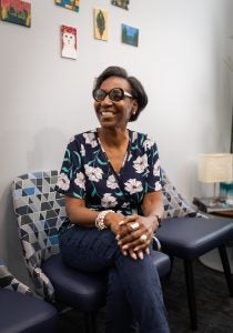 A woman wearing glasses and a patterned shirt smiles on a seat in an office.