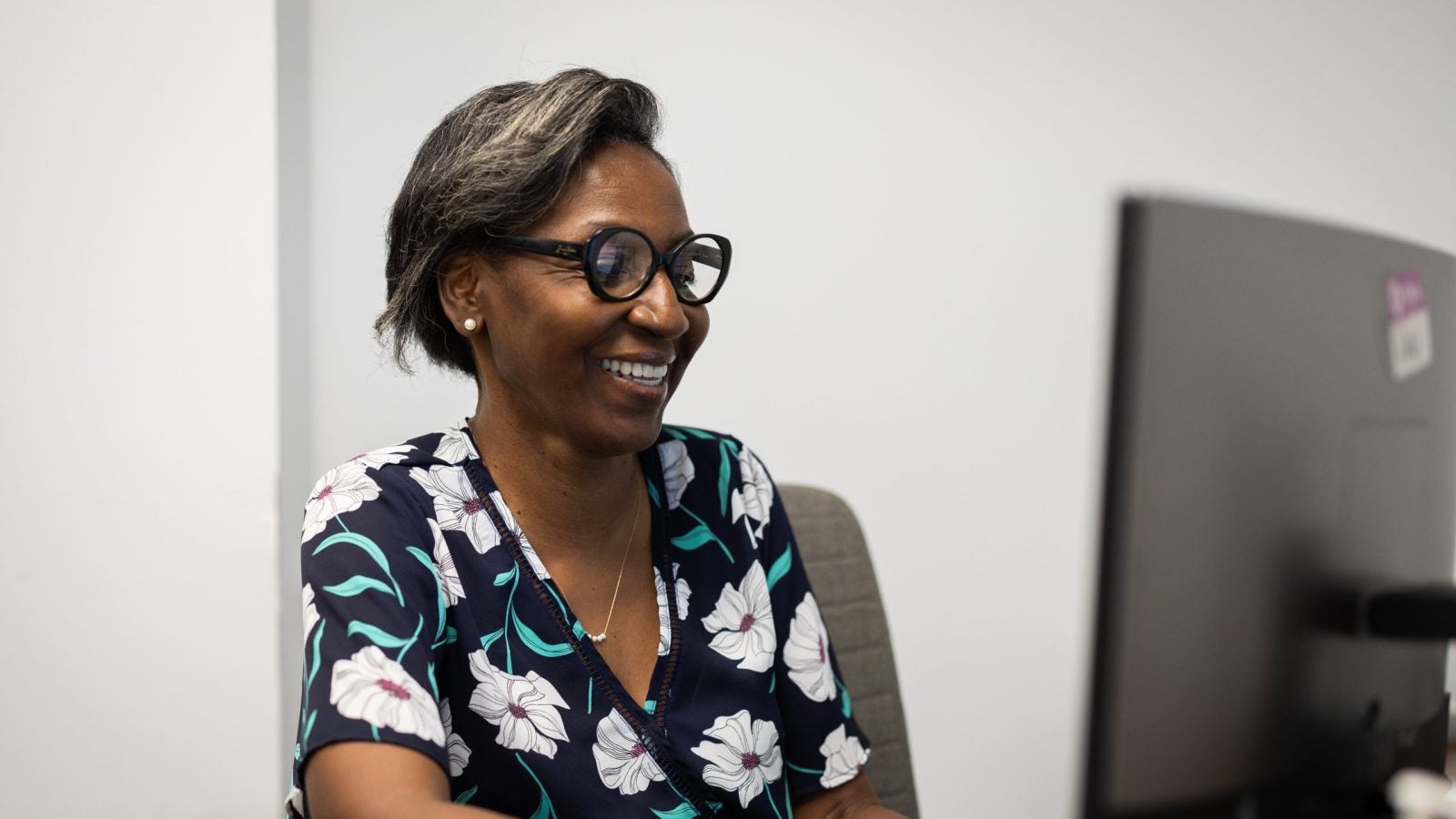 A woman with glasses and a black shirt with white flowers on it smiles as she looks at a computer.