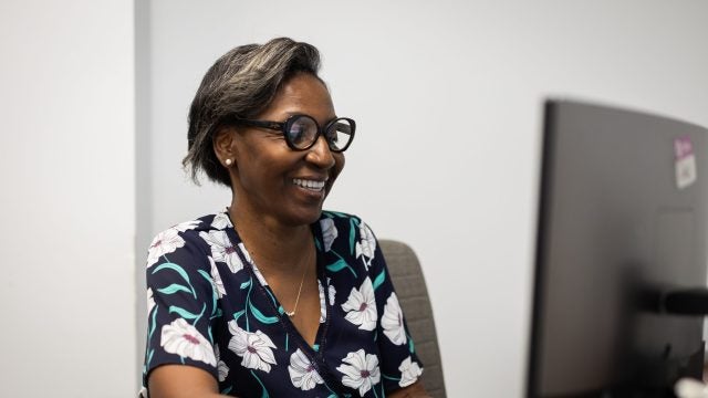 A woman with glasses and a black shirt with white flowers on it smiles as she looks at a computer.