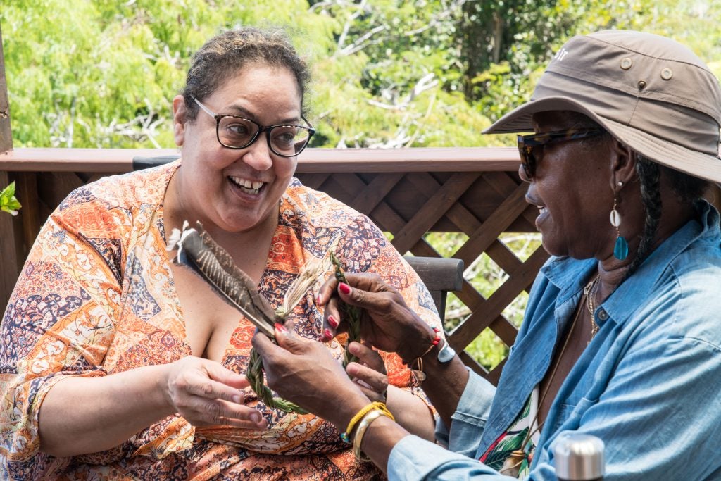 A woman in an orange patterned shirt presents a feather to another woman wearing a hat, sunglasses and a blue shirt