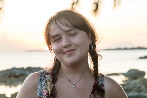 A college student with brown hair in braids smiles in front of a beach sunset.