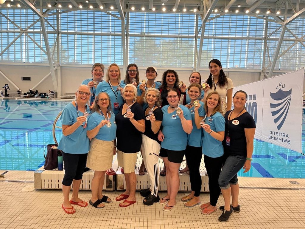 A group of women hold gold medals in front of a pool.