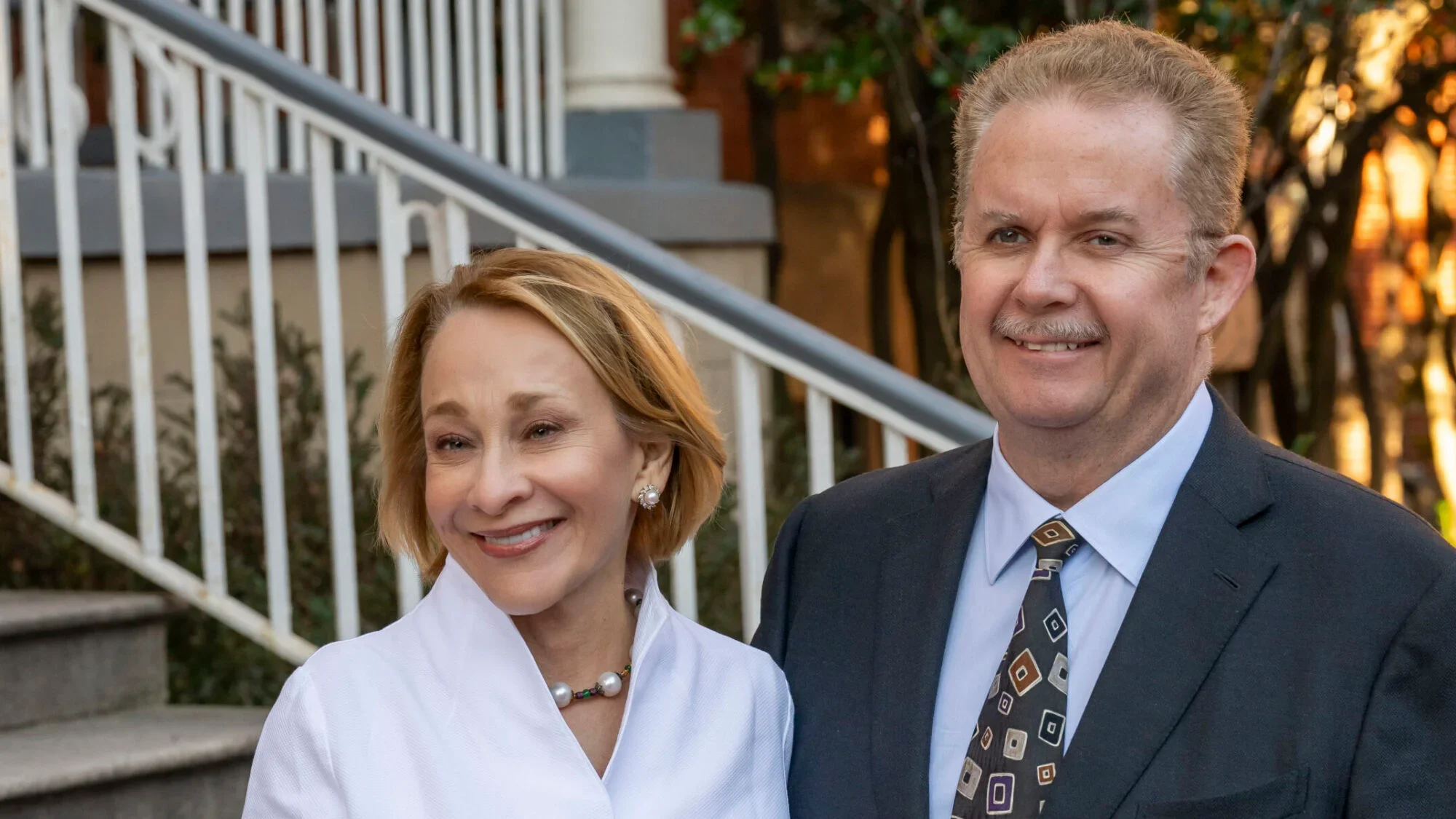 A woman in a white suit and a man in a black suit pose together and smile.