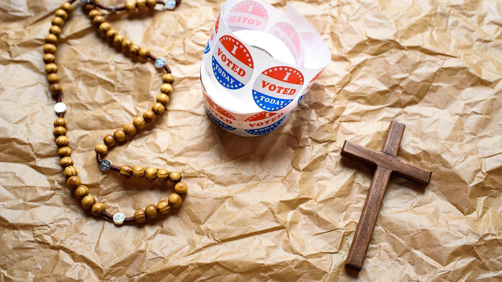 A rosary, a roll of &quot;I voted&quot; stickers, and a wooden cross arranged on a backdrop of crumpled brown paper.