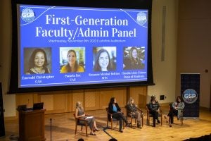 A panel sits on a stage with a projector screen behind them.