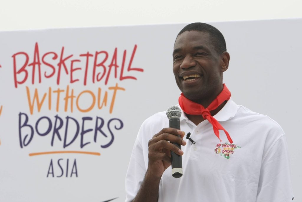 Mutombo in a white shirt in front of a sign for BasketballWithout Borders