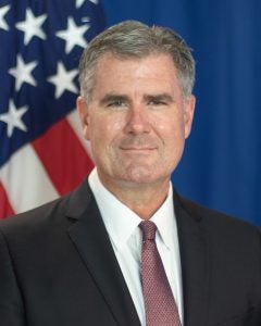 Gillen in business attire with a US flag behind him in an official portrait