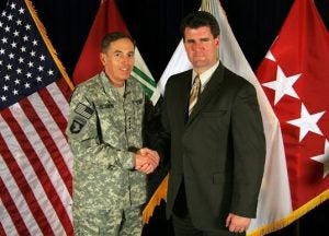 Gillen in a suit shaking hands with David Petraeus in a military uniform in front of flags