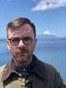 A man with glasses and a jacket stands in front of a blue sea on a sunny day.