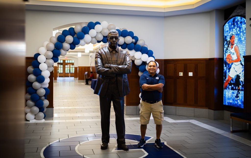 A man in a blue polo shirt stands next to a statue of the late John Thompson Jr. the former coach of the men's basketball team at Georgetown.