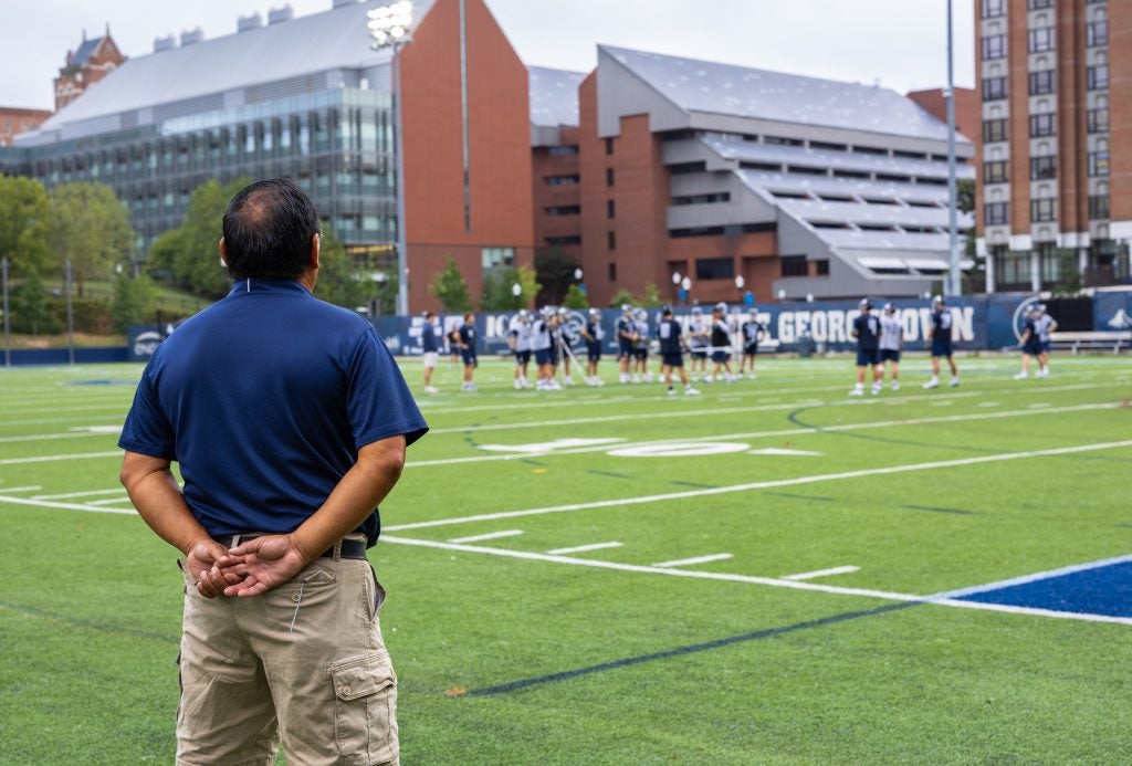 A man in a blue polo shirt and shorts watches lacrosse players practice on a field with his back to the camera and hands behind his back.