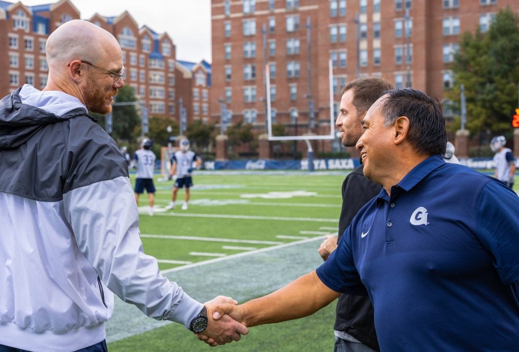 One man in a blue polo shirt shakes hands with another man on the sidelines of a soccer field.