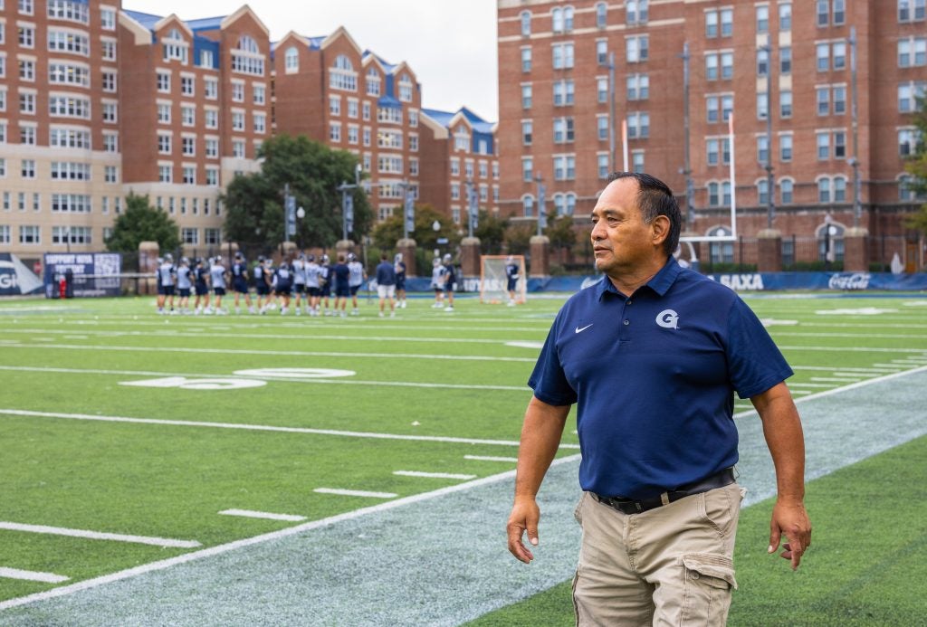 A man in a blue polo shirt walks alongside the sideline of a soccer field.