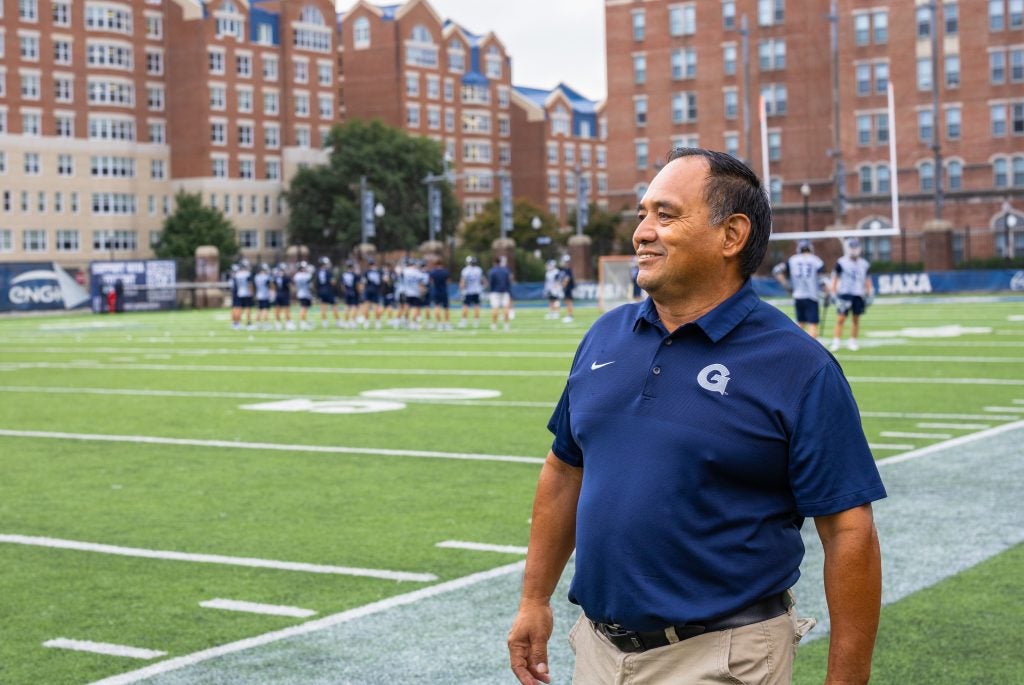 A man in a blue polo shirt walks along the sidelines of a soccer field.