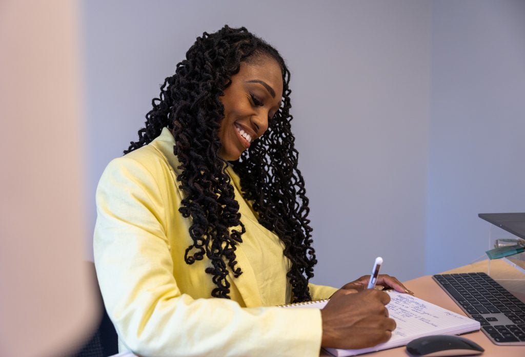 A woman with black hair and a yellow blazer smiles as she writes on a notepad.