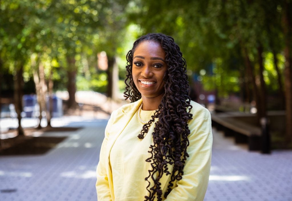 A woman wearing a yellow blazer smiles on a tree-laden pathway at Georgetown.