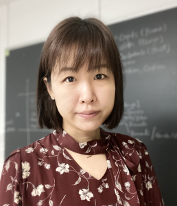 A woman with short brown hair and bangs looks at the camera. A chalkboard is behind her.