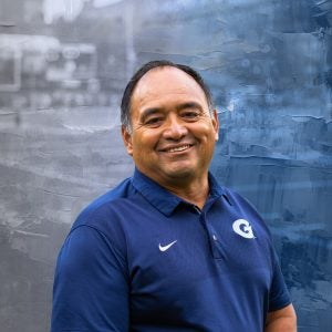 A man in a blue polo shirt with a "G" on it smiles in front of a graphic background.