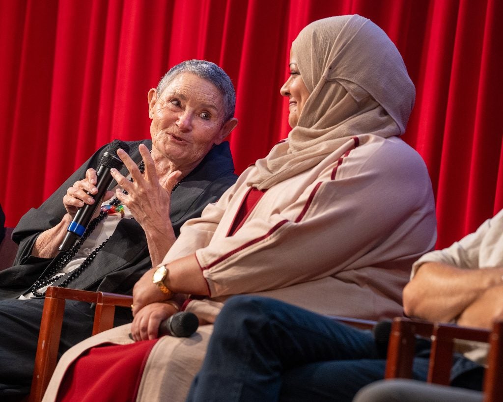 One woman holds a mic and smiles at another woman as they sit talking on stage.