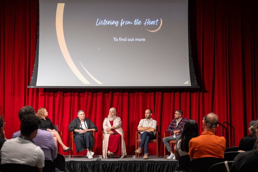 A group of panelists sit on a stage with the screen "Listening from the Heart" behind them.