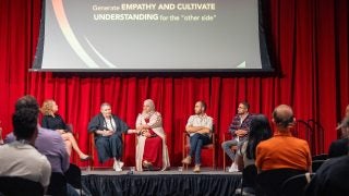 A group of panelists sit on a stage with a screen behind them.