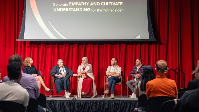 A group of panelists sit on a stage with a screen behind them.